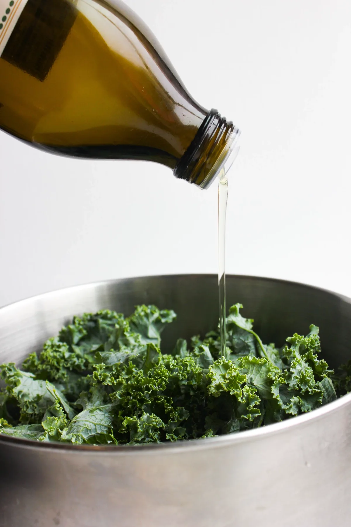 Extra virgin olive oil being drizzled from a bottle over fresh kale leaves in a bowl